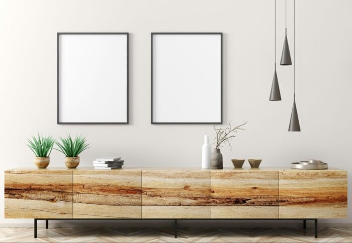 Minimalist living room with a wooden sideboard, two blank frames, plants, and decorative items. Sleek pendant lights hang above, conveying a modern tone.