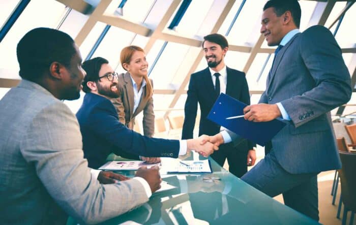 a group of five professionals in formal attire engage in a positive business meeting, smiling as two of them shake hands across a glass table