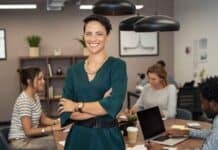 Do You Have a Marketing Strategy for the Next Quarter? Smiling woman in a green dress stands confidently with arms crossed in an office. Three colleagues work at a table in the background, creating a collaborative and positive atmosphere.