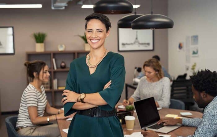 Smiling woman in a green dress stands confidently with arms crossed in an office. Three colleagues work at a table in the background, creating a collaborative and positive atmosphere.