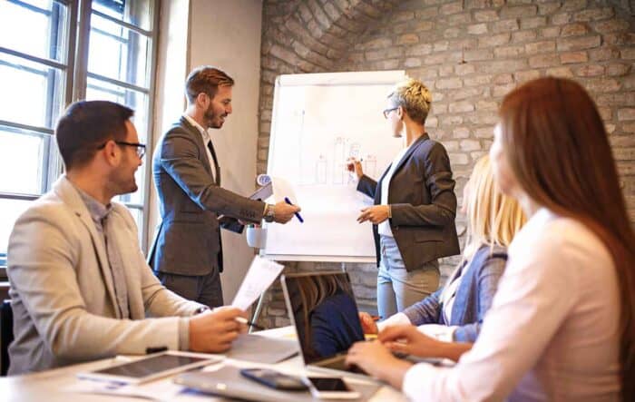 A group of business professionals in a meeting room, discussing at a whiteboard. The mood is collaborative, with natural light from large windows.