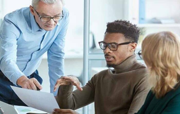 Three colleagues collaborate in an office. A senior man points at a document while two younger individuals, one with glasses, listen attentively.