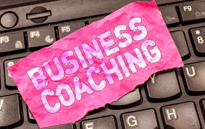 pink paper with "Business Coaching" in bold white letters placed on a black keyboard. The tone is professional and motivational