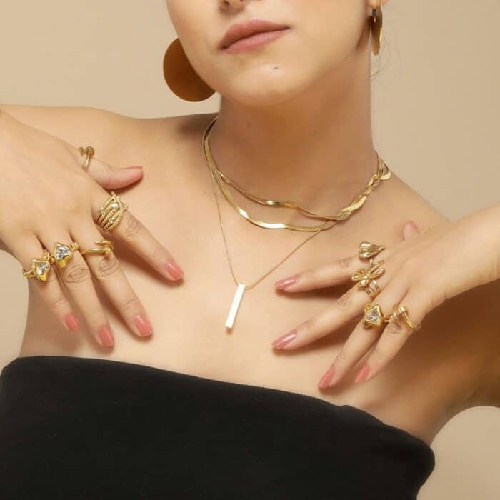 A woman displays elegant gold jewelry, including layered necklaces, multiple rings, and hoop earrings. She wears a strapless black top, conveying a chic and sophisticated vibe.