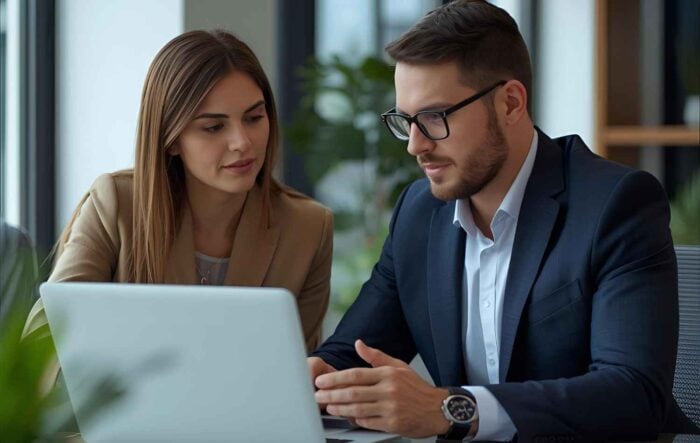 A man and woman in business attire are focused on a laptop in a modern office with plants in the background, conveying collaboration and concentration.