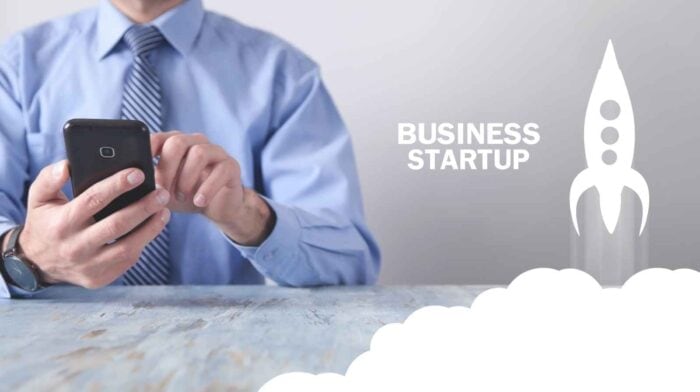a man in a blue shirt and tie uses a smartphone at a wooden table. Beside him, the text "Business Startup" and a rocket icon suggest innovation