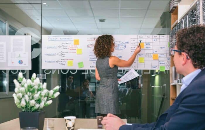 a woman presents at a meeting, pointing to a project management chart on a glass wall. Colleagues listen attentively. The setting is professional and focused