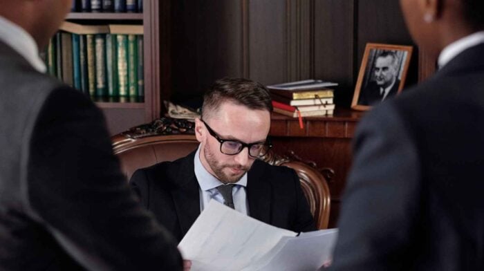 A serious man in a business suit with glasses reviews documents in an office with bookshelves. Two clients stand facing him, and a framed photo is in the background