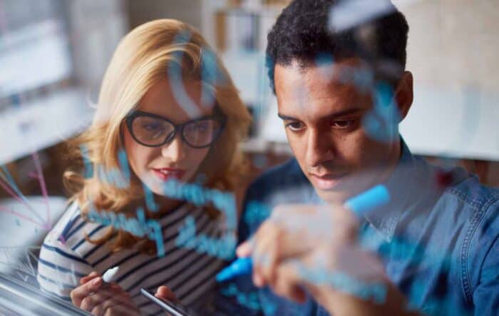 Two people focus intently while writing with markers on a glass board, brainstorming ideas. The image conveys collaboration and creativity.