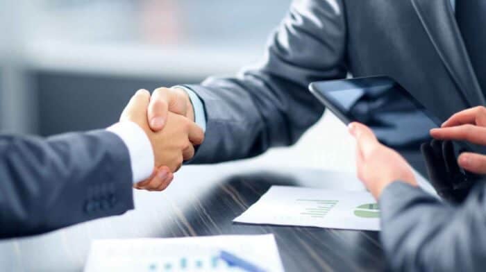 two people in formal suits are shaking hands over a table, signifying a business agreement. One holds a tablet, and papers with charts are visible
