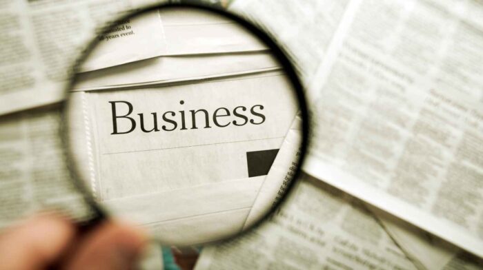 a magnifying glass focuses on the word "Business" in a newspaper, suggesting analysis or investigation. The background shows blurred text, conveying a professional tone