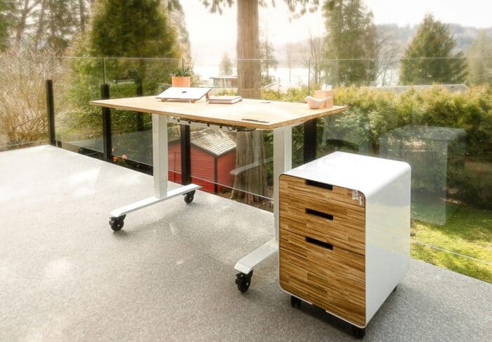 Modern outdoor workspace with a sleek wooden desk and rolling storage unit on a patio. Background features lush greenery and a serene lake view.