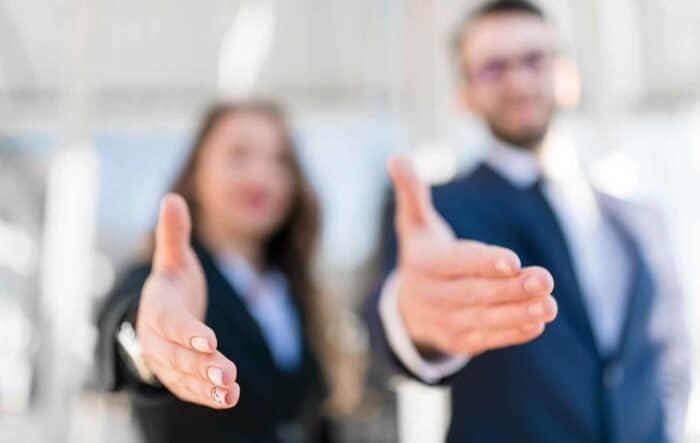 Two people in business attire extend hands forward for a handshake, symbolizing welcome or agreement. The background is softly blurred, creating a professional tone.