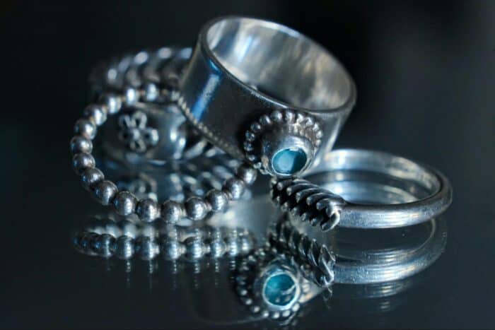 A stack of silver rings, including a thick band with a green gemstone and a beaded ring, reflecting on a shiny black surface, evokes elegance.