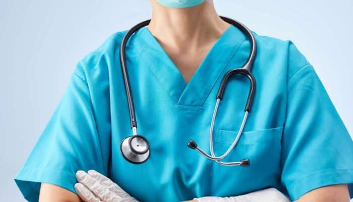 A medical professional in blue scrubs with a stethoscope around their neck stands confidently, wearing gloves and a mask, background softly blurred.