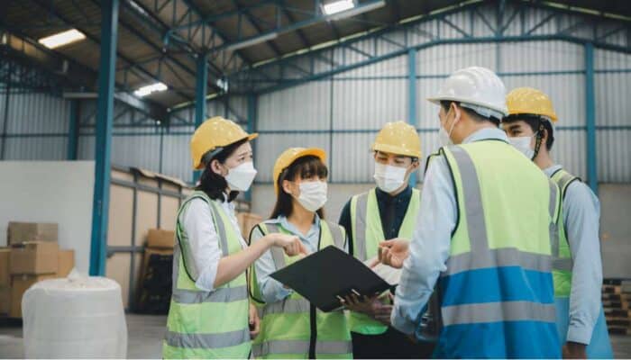 A group of workers in hard hats and reflective vests stand in a warehouse, discussing a document. They appear focused and collaborative, conveying teamwork.