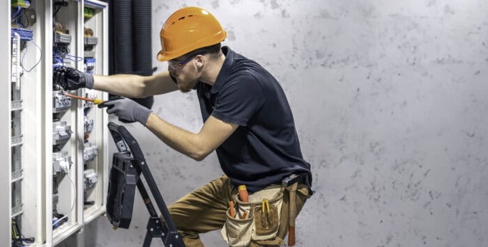 Electrician wearing an orange hard hat and tool belt works on a circuit panel. He stands on a ladder with tools, focused and attentive. Industrial setting.