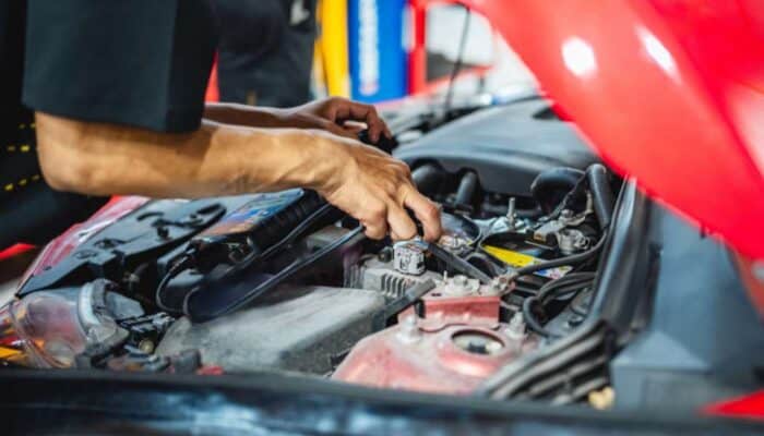 Mechanic's hands working on a car engine under an open red hood. Various tools and parts are visible, conveying a sense of precision and focus.