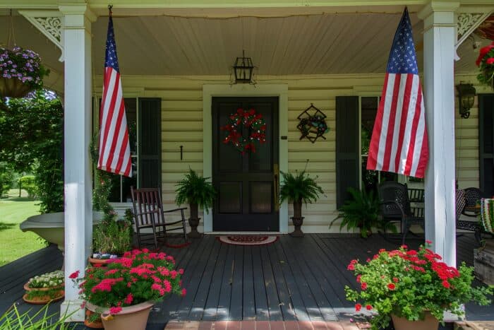 Front porch with American flags, a dark door adorned with a red wreath, and potted red flowers. Rocking chair and black chairs suggest a cozy, patriotic atmosphere.