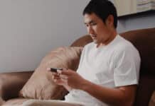 The Card Effect: How Playing Solitaire Can Actually Enhance Your Life A man in a white shirt sits on a brown sofa, focused on his smartphone. The room is softly lit, creating a calm and relaxed atmosphere.