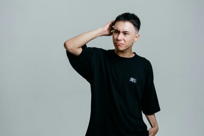 A person in a black T-shirt stands against a grey background, scratching their head with a puzzled expression, conveying confusion or contemplation.