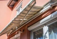 The Ultimate Spring Hosting Hack: Why Your Deck Needs a Roof on Demand Sunlit image of a tan retractable awning over a window on a peach-colored house exterior. The awning casts shadows, creating a warm, summery atmosphere.