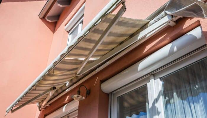 Sunlit image of a tan retractable awning over a window on a peach-colored house exterior. The awning casts shadows, creating a warm, summery atmosphere.