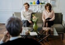 How Counseling Can Protect Your Family During a Divorce A couple sits apart on a sofa in a therapy session, looking tense. A therapist observes. A flower vase and notebook on the table add to the formal setting.