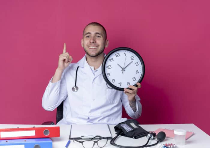 Smiling doctor in a white coat holds a clock, emphasizing time, against a pink background. Office supplies and medical tools are on the desk.