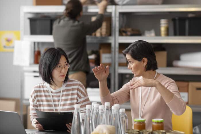 Two women sit at a table discussing, surrounded by bottles and jars, with a clipboard in hand. A person in the background organizes shelves. The setting is collaborative and busy.
