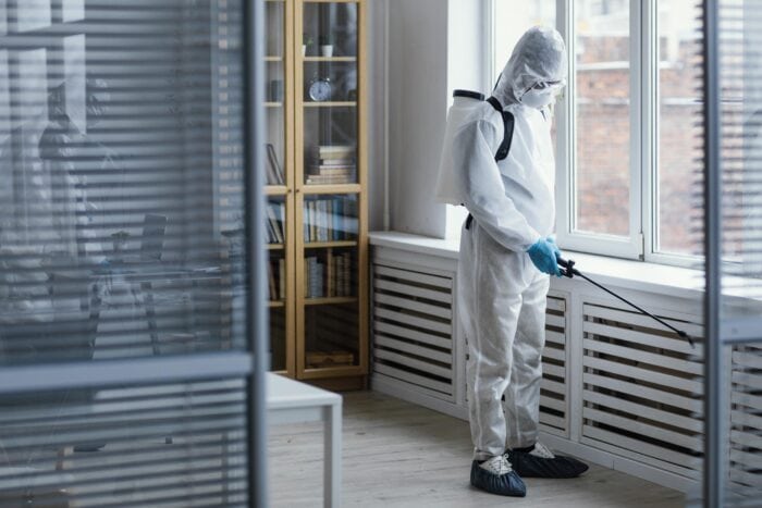 Person in protective suit and mask, disinfecting a bright room beside large windows. Cabinets with books visible, setting a serious, sterile tone.