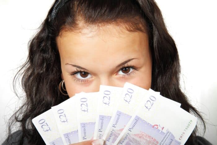Young woman with dark hair playfully holds several British banknotes in front of her face, eyes visible above, conveying a sense of mystery and excitement.
