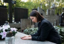 Finding Space to Grieve: Managing Sudden Funeral Expenses Without the Financial Panic A woman in a black coat leans over a grave with a solemn expression, surrounded by headstones and greenery. Pink and white flowers adorn the grave.