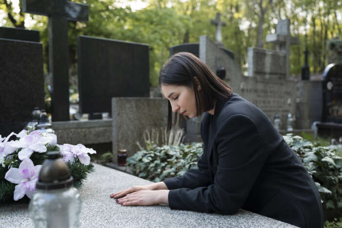 A woman in a black coat leans over a grave with a solemn expression, surrounded by headstones and greenery. Pink and white flowers adorn the grave.