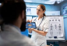 Surviving the Summer Rush: How Digital Interfaces Save the Hospitality Industry A female doctor in a lab coat holds a tablet displaying medical images, explaining to a colleague. A human skeleton chart is visible in the background.