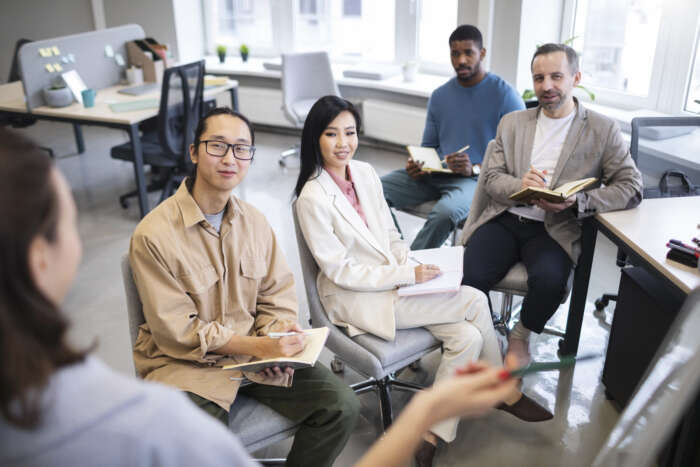 A small group of professionals seated in a modern office, listening attentively and taking notes during a meeting or training session led by a speaker in the foreground.