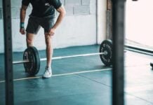Building Muscle vs. Shedding Pounds: How Your Workout Should Match Your Goal A man in athletic gear prepares to lift a barbell in a gym. He is focused, wearing a black shirt and shorts. The space is industrial with ample light.