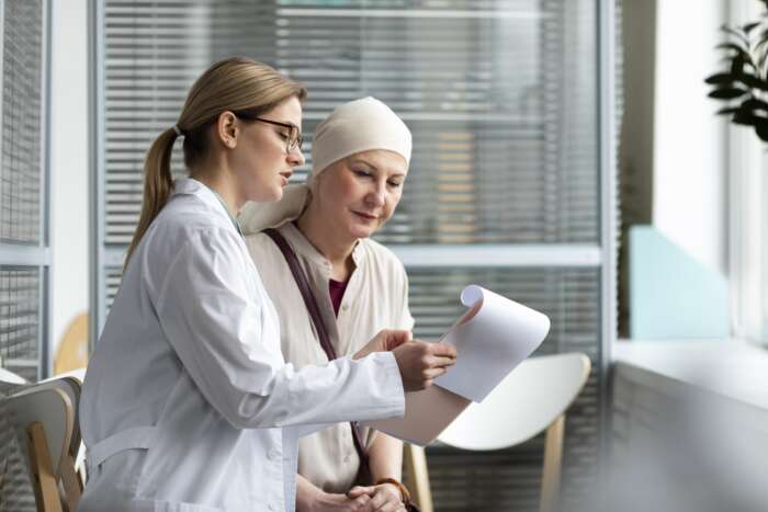 A female doctor in a white coat explains medical documents to a patient wearing a headscarf, in a bright clinic. The atmosphere is calm and supportive.