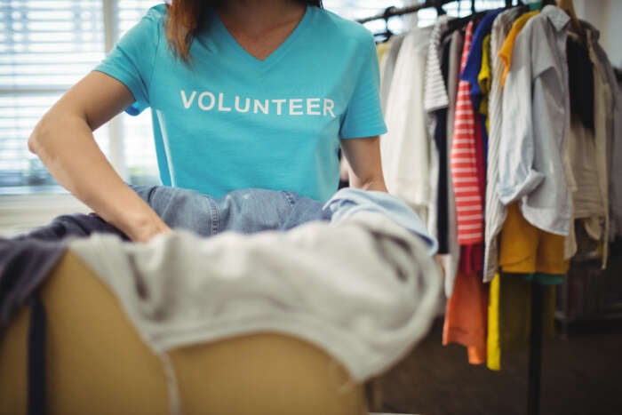 A person wearing a blue "Volunteer" shirt folds clothes. In the background, a rack with assorted colorful garments is visible, conveying community service.