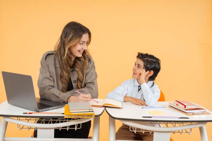 A woman and a young boy sit at desks with notebooks and a laptop, smiling at each other, against a plain orange background.