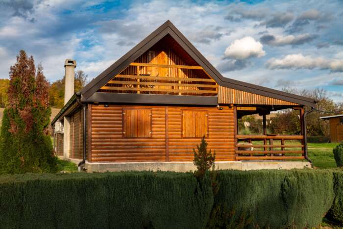 A cozy wooden cabin with closed shutters and a sloped roof sits in a lush, green landscape under a partly cloudy sky, evoking tranquility and warmth.
