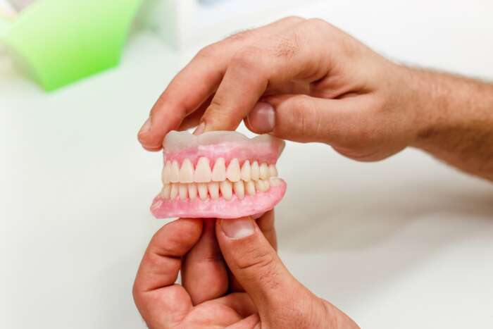 Two hands holding a set of pink and white dental dentures on a bright, clean surface, conveying a clinical and educational tone.