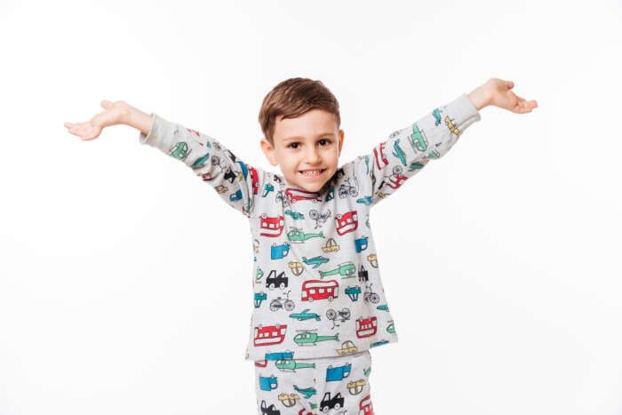 Young boy smiling with his arms outstretched against a white background. He's wearing pajamas featuring playful car and animal patterns, conveying joy.