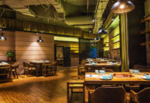 Lunch Break: Why Hiring a Local Restaurant for Your Next Group Lunch is the Best Move Warmly lit rustic restaurant interior with wooden tables, chairs, pendant lights, exposed ceiling, plants, and cozy amber-toned decor.