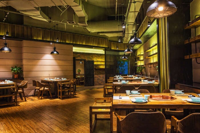 Warmly lit rustic restaurant interior with wooden tables, chairs, pendant lights, exposed ceiling, plants, and cozy amber-toned decor.