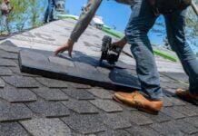 A Seasonal Replacement: Why Fall is the Perfect Season to Replace Your Roof Roofing worker installs gray asphalt shingles on a steep house roof, using a nail gun under bright blue skies; hands-on construction scene.