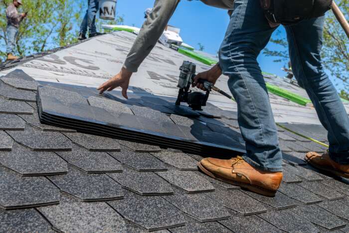 Roofing worker installs gray asphalt shingles on a steep house roof, using a nail gun under bright blue skies; hands-on construction scene.