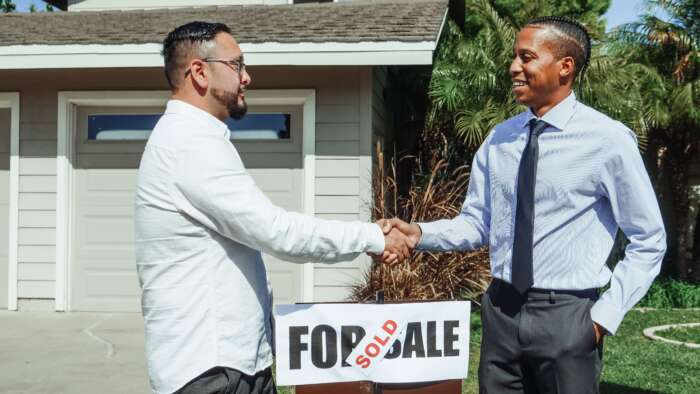 Two men shake hands outside a house beside a “SOLD” real estate sign, suggesting a successful sale in a bright, friendly outdoor scene.
